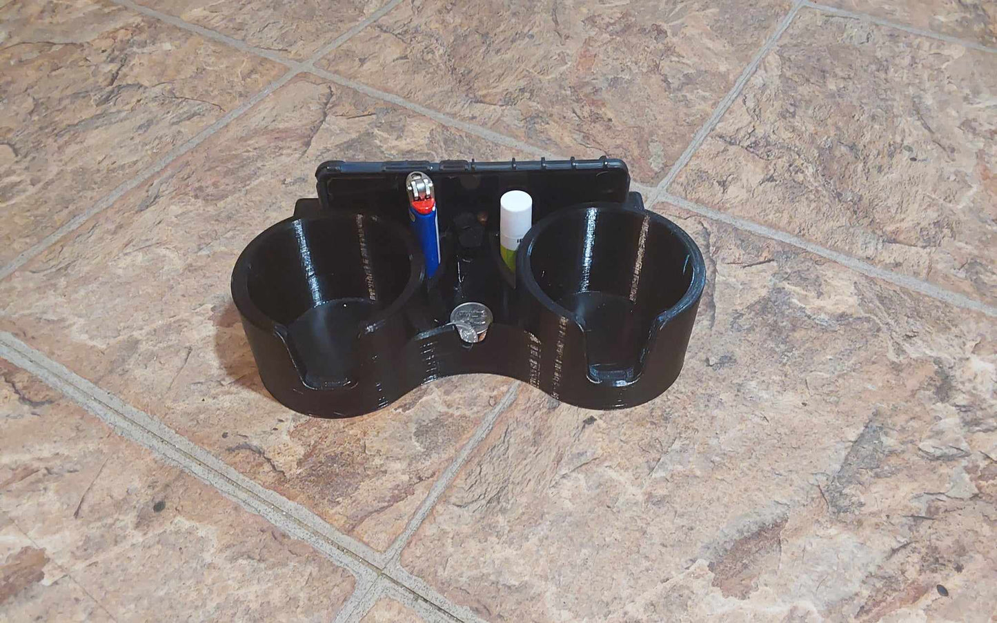 A black dual cup holder attachment with a slot for a phone or ipad, featuring two cup holders and a small catch-all dish, placed on a tiled surface.