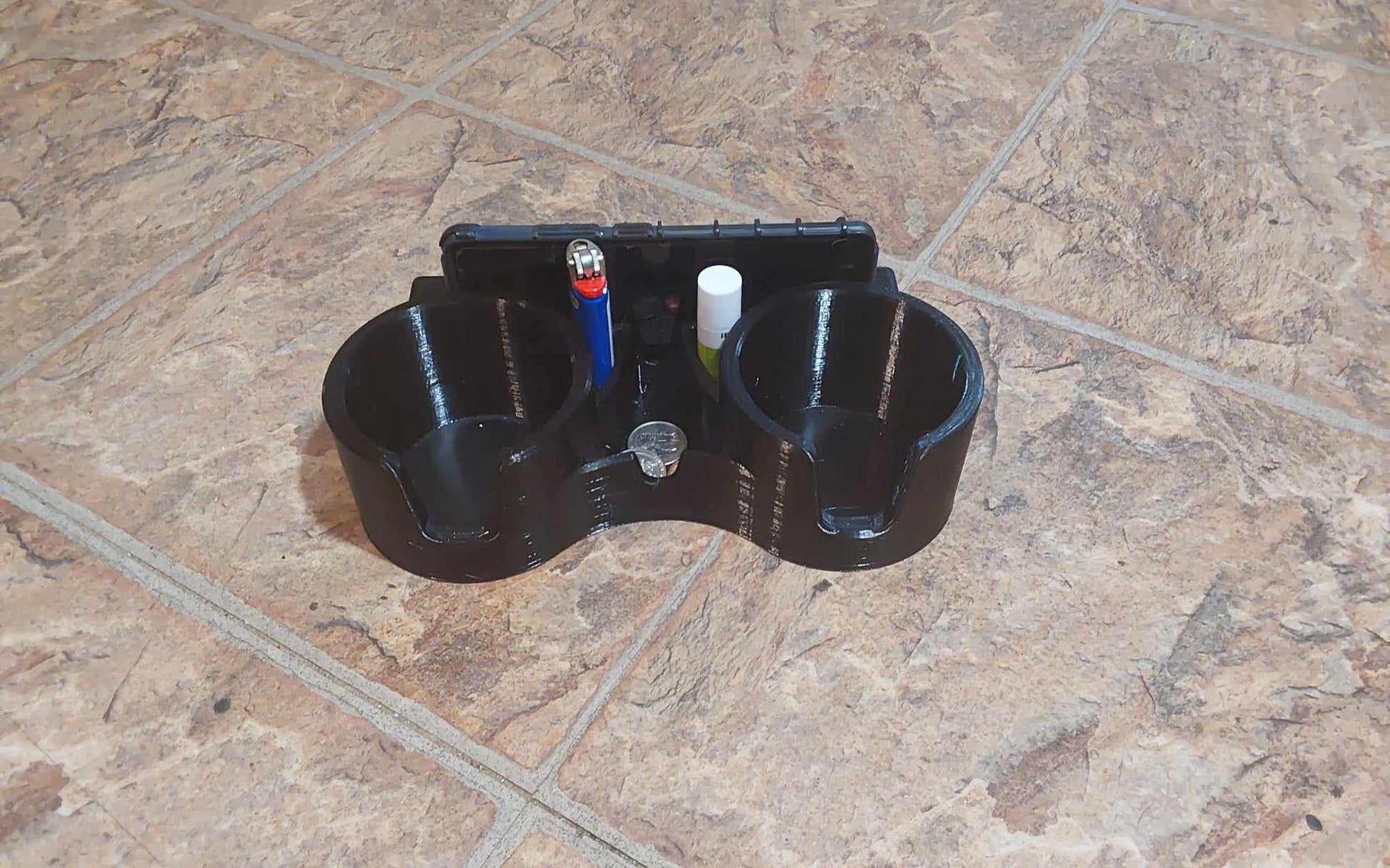 A black dual cup holder attachment with a slot for a phone or ipad, featuring two cup holders and a small catch-all dish, placed on a tiled surface.